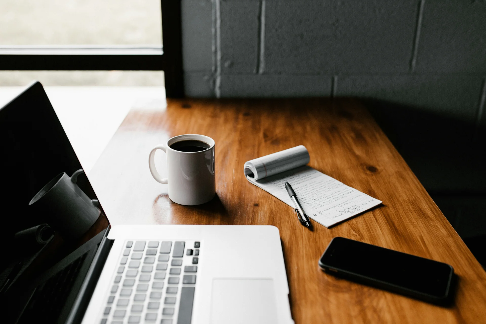 een bureau met een laptop, een tas koffie, een smartphone en een notitieblokje 